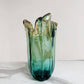 Die mittelgroße Murano-Dream-Vase „Venezia“ aus grünem Murano-Glas (H 25, Ø 14 cm) mit gewelltem Rand und sanftem Farbverlauf von Grün bis Blaugrün steht auf weißem Marmor vor hellem Hintergrund.