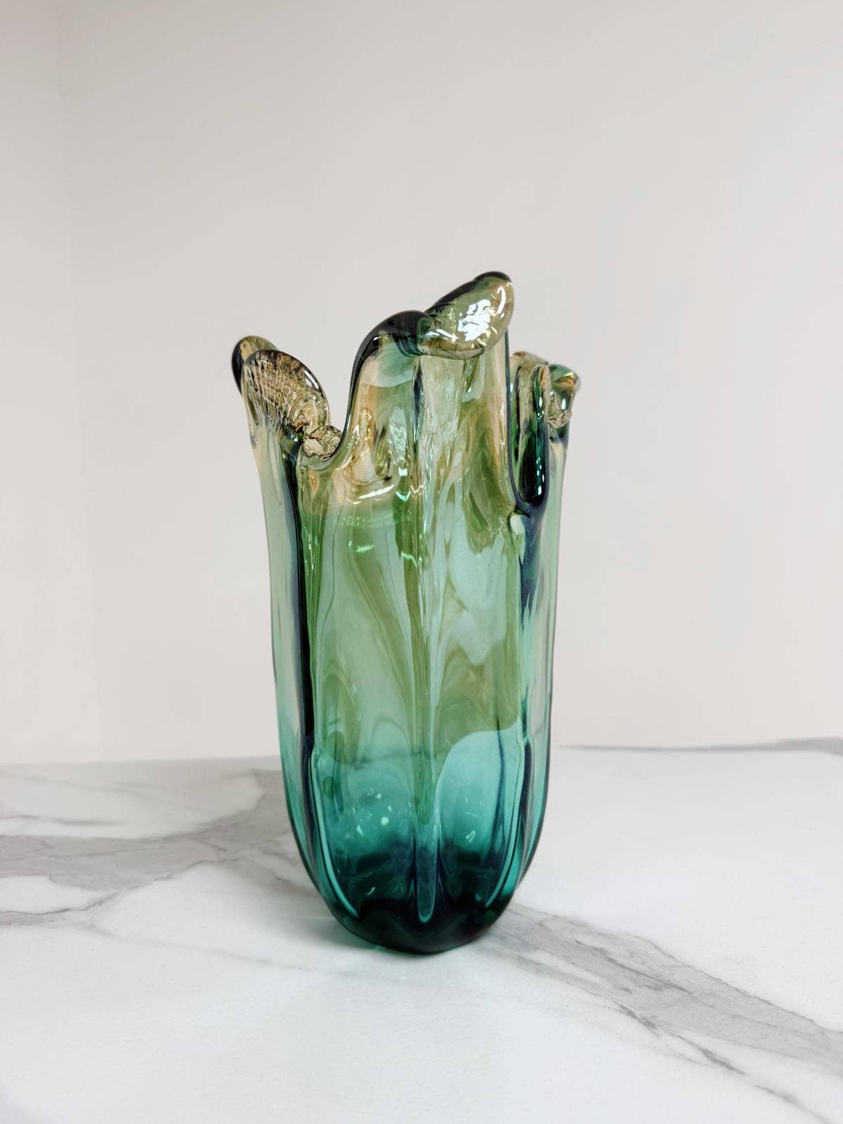 Die mittelgroße Murano-Dream-Vase „Venezia“ aus grünem Murano-Glas (H 25, Ø 14 cm) mit gewelltem Rand und sanftem Farbverlauf von Grün bis Blaugrün steht auf weißem Marmor vor hellem Hintergrund.