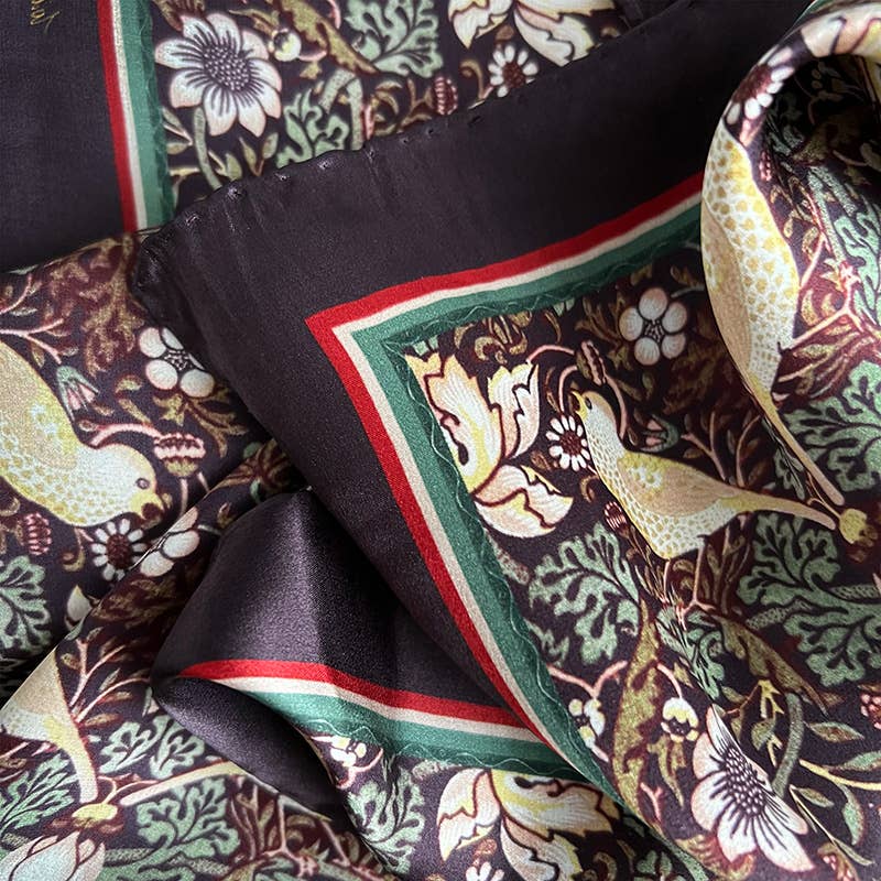 Eine Nahaufnahme des Koustrupco | Seidenschal Morris Strawberry Thief Bordeaux (50 x 50 cm) von Koustrupco mit detaillierten Blumen- und Vogelmustern in Gelb, Grün und Creme, umrandet mit roten und grünen Streifen. Die Seide wirkt weich und sanft gefaltet.