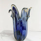 Die Murano Dream | Die große Giusy-Vase aus originalem blauem Murano-Glas (H 32, Ø 20 cm) besticht durch ihre wellenförmigen Ränder und klaren Glaselemente – ein traditionelles italienisches Meisterwerk, präsentiert auf weißem Marmor.