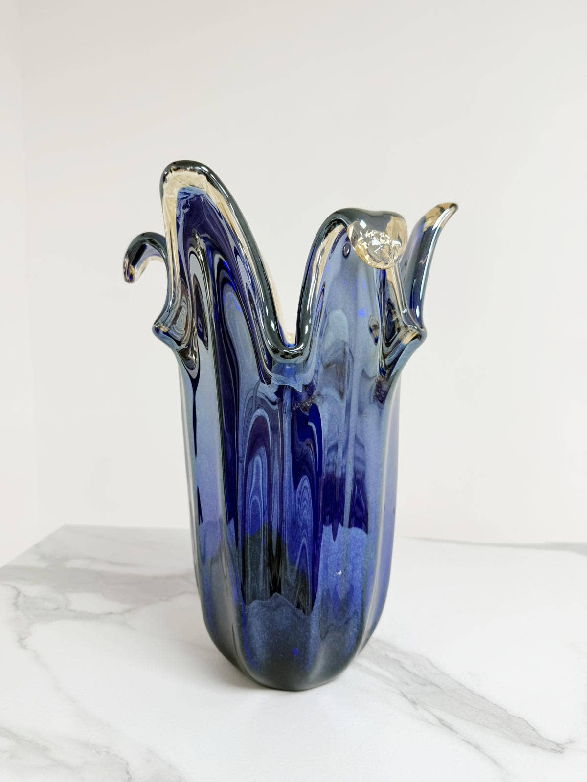 Die Murano Dream | Die große Giusy-Vase aus originalem blauem Murano-Glas (H 32, Ø 20 cm) besticht durch ihre wellenförmigen Ränder und klaren Glaselemente – ein traditionelles italienisches Meisterwerk, präsentiert auf weißem Marmor.