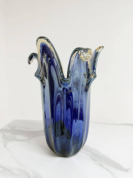Die Murano Dream | Die große Giusy-Vase aus originalem blauem Murano-Glas (H 32, Ø 20 cm) besticht durch ihre wellenförmigen Ränder und klaren Glaselemente – ein traditionelles italienisches Meisterwerk, präsentiert auf weißem Marmor.