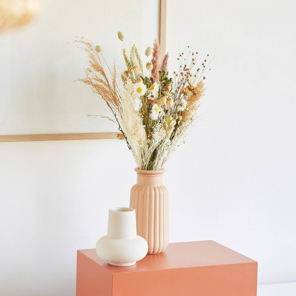 Ein mittelgroßer „Getrockneter Feldblumenstrauß ‚Natur‘“ von Wildflowers by Floriette steht in einer beigen Vase auf einem korallenfarbenen Tisch neben einer kleinen weißen Vase. Im Hintergrund sind eine weiße Wand und ein Kunstwerk in einem hellen Holzrahmen zu sehen, das teilweise sichtbar ist.