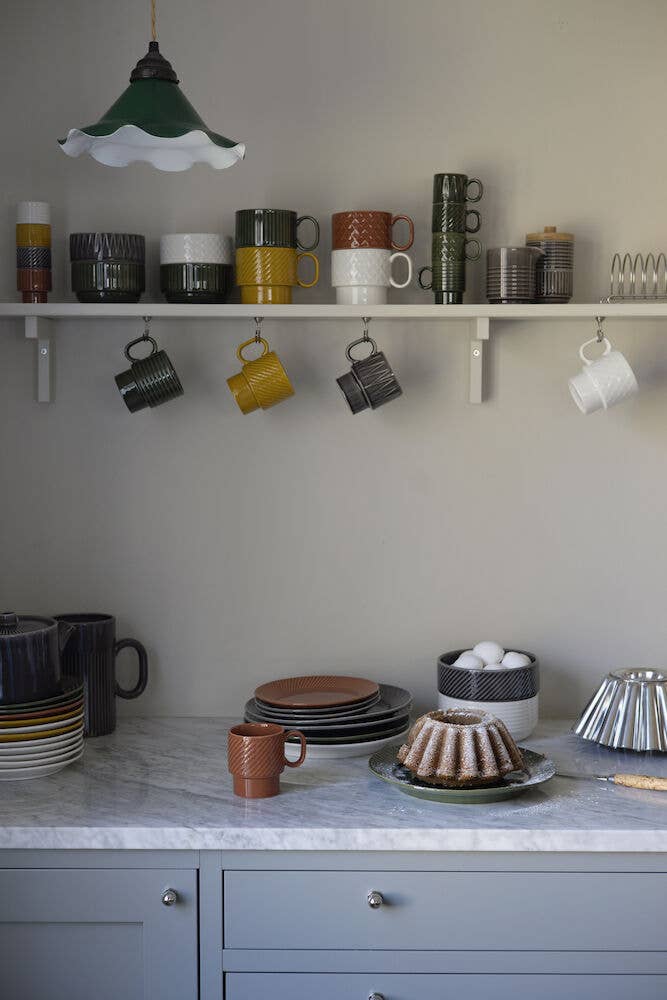 Eine Küchenszene mit einer Arbeitsplatte aus Marmor, gestapeltem Geschirr, Tassen, Kuchen und Küchenutensilien. Auf dem Wandregal unter einer grünen Pendelleuchte steht das Sagaform GmbH Kaffeebecher Gelb 2-teilig Set und sorgt für skandinavisches Design-Flair.