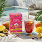 Eine Schachtel English Tea Shop | 'Mango Splash' BIO 20 Teebeutel steht auf einem Tisch neben einem Glas exotischen Eistees mit Früchten. Aufgeschnittene Mango, Ananas, ein Pool, ein Stuhl und Pflanzen ergänzen diese sonnige Umgebung im Freien.