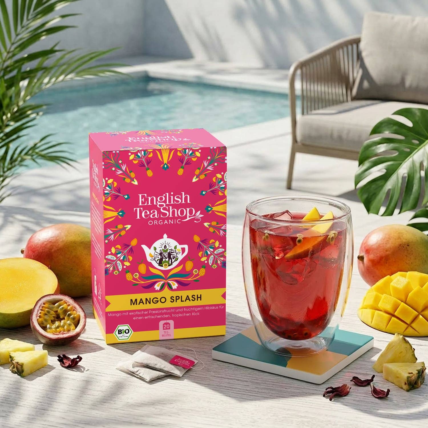 Eine Schachtel English Tea Shop | 'Mango Splash' BIO 20 Teebeutel steht auf einem Tisch neben einem Glas exotischen Eistees mit Früchten. Aufgeschnittene Mango, Ananas, ein Pool, ein Stuhl und Pflanzen ergänzen diese sonnige Umgebung im Freien.