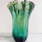 Die Murano Dream „Fazzoletto“-Vase aus grünem Murano-Glas (H 32 Ø 24 cm) besticht durch ihre hohe, wellenförmige Form und den sanften Farbverlauf von Grün zu Transparent – handgefertigt und stilvoll vor hellgrauem Hintergrund präsentiert.