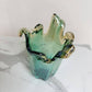 Die mittelgroße Vase „Venezia“ von Murano Dream aus grünem Murano-Glas (H 25, Ø 14 cm) mit welligen, organisch geformten Rändern steht auf weißem Marmor vor einem schlichten Hintergrund.