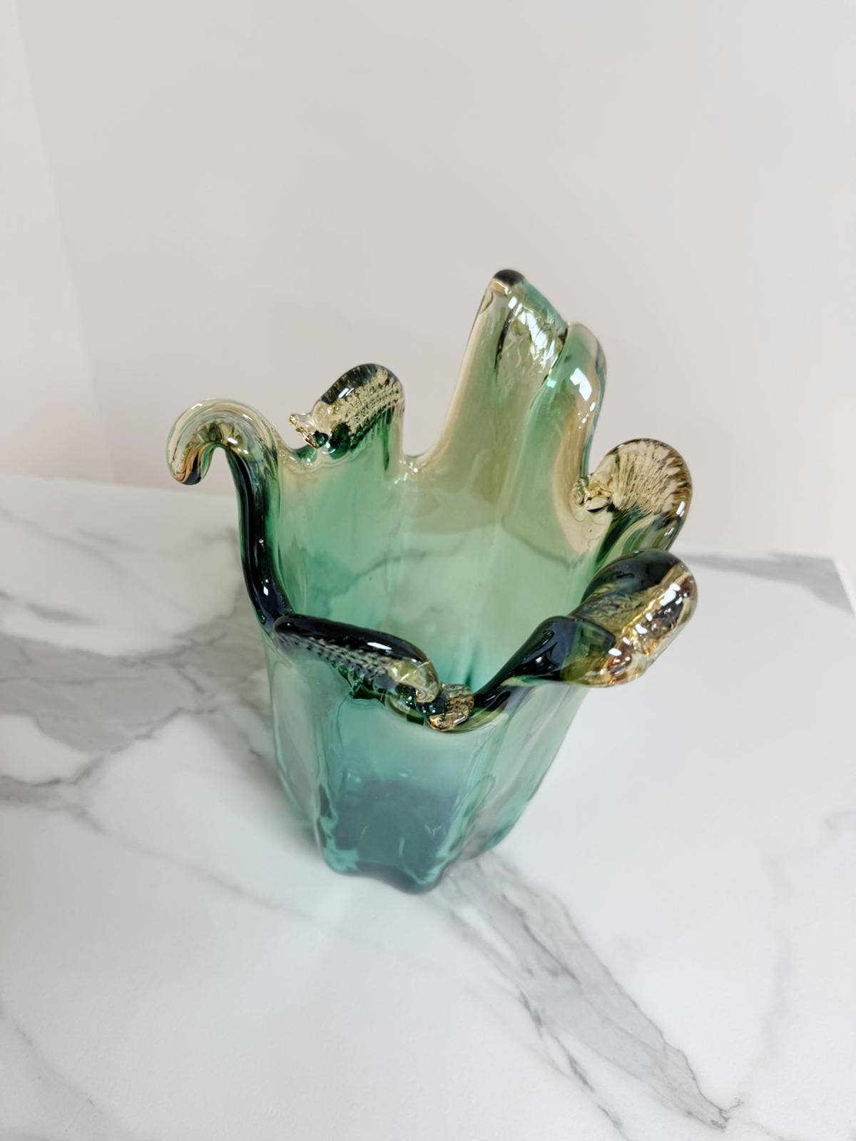 Die mittelgroße Vase „Venezia“ von Murano Dream aus grünem Murano-Glas (H 25, Ø 14 cm) mit welligen, organisch geformten Rändern steht auf weißem Marmor vor einem schlichten Hintergrund.