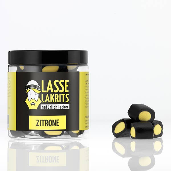 Eine LASSE LAKRITS Dose Lakritz Zitrone sitzt neben schwedischen Lakritzstücken mit Zitronenkern, mit einem Cartoon-Gesicht und einem auffälligen schwarz-gelben Branding auf einer weißen Dose.