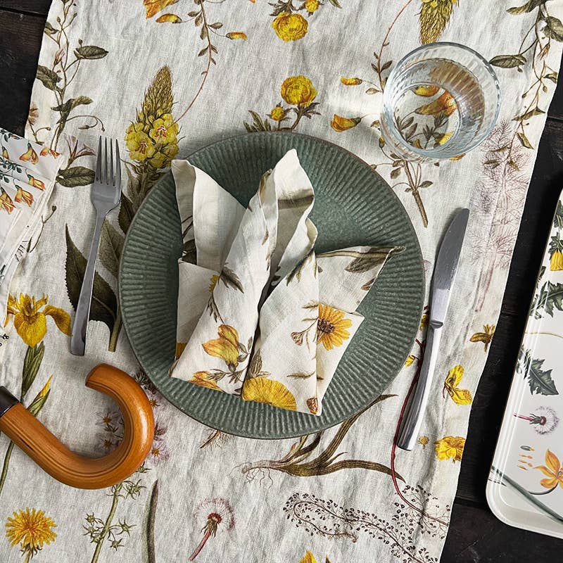 Auf einer Tischdecke mit botanischem Muster steht ein grüner Teller mit einer Koustrupco-Leinenserviette mit gelbem Blütenmotiv (45 x 45 cm). Links und rechts vom Teller liegen eine Gabel und ein Messer, darüber steht ein Wasserglas, und gelbe Blüten unterstreichen die botanische Tischdekoration.