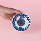 Eine Hand hält die „Alfar Tierra Cocida Kleine Keramik Knoblauchreibeplatte ‚Nerja‘“ – eine runde, weiße Keramikplatte mit traditionellen blauen Blattmustern und einer strukturierten Spirale in der Mitte vor einem rosa Hintergrund.