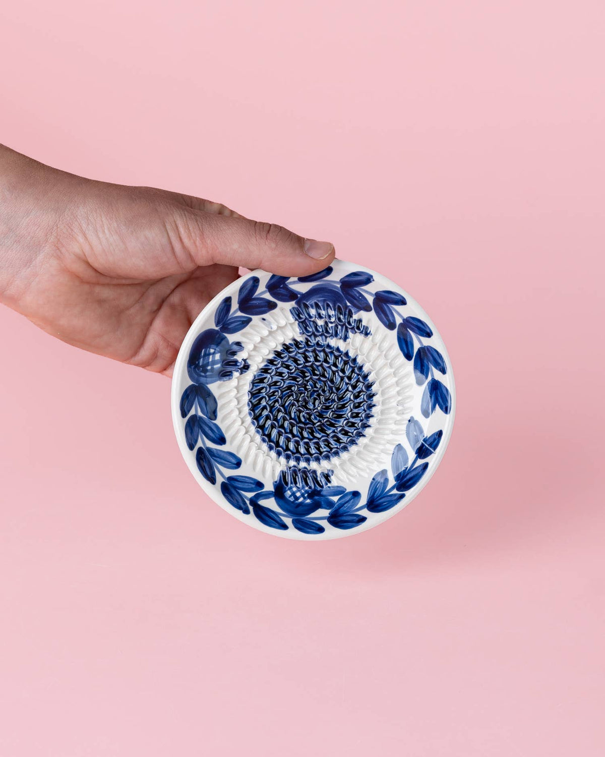 Eine Hand hält die „Alfar Tierra Cocida Kleine Keramik Knoblauchreibeplatte ‚Nerja‘“ – eine runde, weiße Keramikplatte mit traditionellen blauen Blattmustern und einer strukturierten Spirale in der Mitte vor einem rosa Hintergrund.