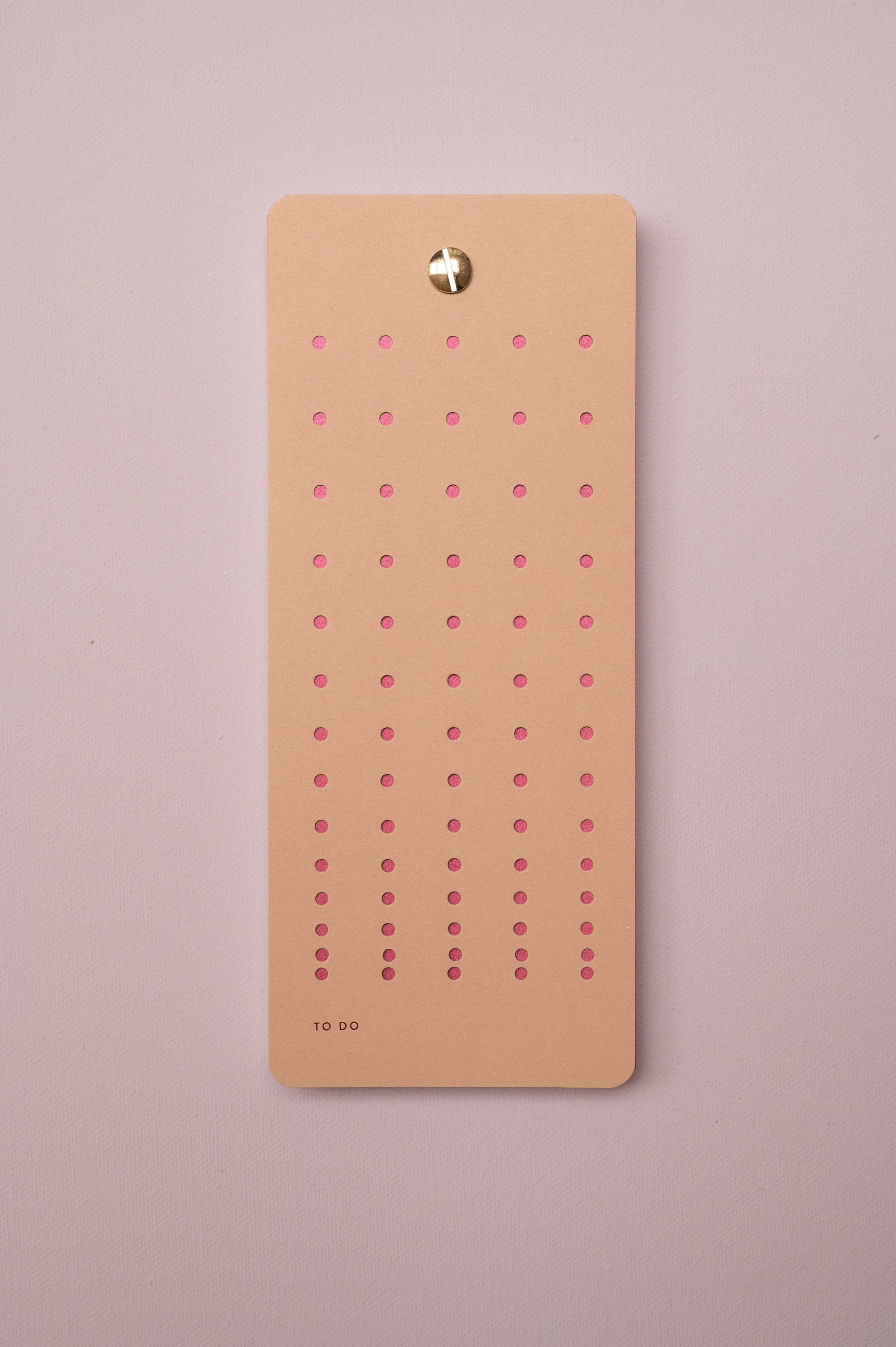 Die Aufgabenliste 'Nougat' von Tinne Mia aus FSC-zertifiziertem, nachhaltigem Papier mit fünf Reihen pinker Punkte ist per goldener Pinnnadel an einer rosa Wand befestigt. Unten links steht "TO DO" in kleiner Schrift.