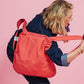 Eine Person in einem Outfit aus dunklem Denim trägt eine große, rot karierte Notabag | Wandelbare Tasche und Rucksack „Beeren“ über der Schulter und steht vor einem rosa Hintergrund. Diese stilvolle Notabag von Notabag dient sowohl als Einkaufstasche als auch als Rucksack und bietet so Vielseitigkeit für jeden Anlass.