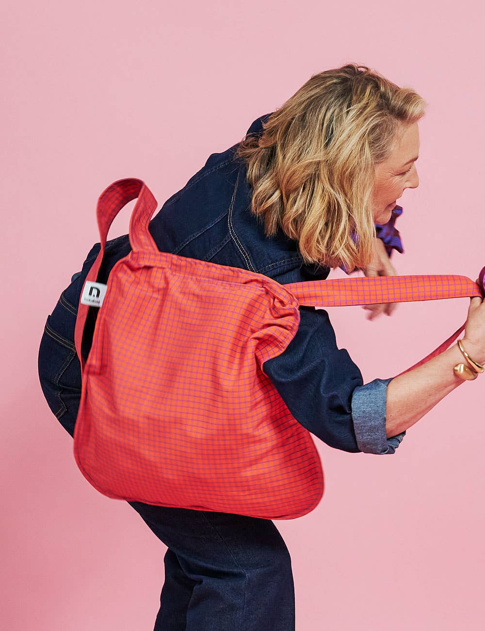 Eine Person in einem Outfit aus dunklem Denim trägt eine große, rot karierte Notabag | Wandelbare Tasche und Rucksack „Beeren“ über der Schulter und steht vor einem rosa Hintergrund. Diese stilvolle Notabag von Notabag dient sowohl als Einkaufstasche als auch als Rucksack und bietet so Vielseitigkeit für jeden Anlass.