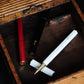 Drei ENTREPÔT YSTUDIO Harz-Rollerball-Stifte (rot, schwarz, weiß) mit Goldspitzen liegen diagonal in einer rustikalen Vintage-Holzbox mit Schiebedeckel. Das sechseckige Design der Stifte verleiht der offenen Auslage Eleganz.