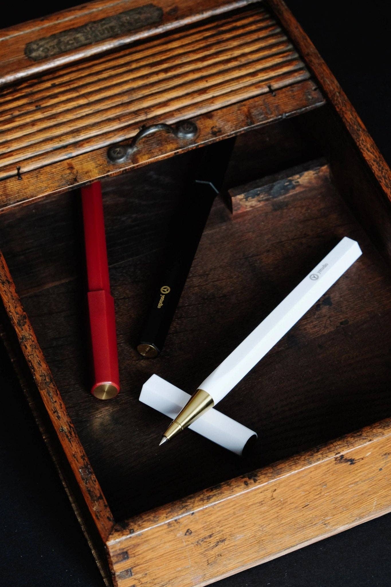 Drei ENTREPÔT YSTUDIO Harz-Rollerball-Stifte (rot, schwarz, weiß) mit Goldspitzen liegen diagonal in einer rustikalen Vintage-Holzbox mit Schiebedeckel. Das sechseckige Design der Stifte verleiht der offenen Auslage Eleganz.