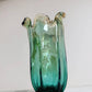 Eine mittelgroße, grüne Murano-Glasvase „Venezia“ von Murano Dream (H 25 cm, Ø 14 cm) mit gewelltem Rand und dezenten goldenen Akzenten steht auf weißem Untergrund vor einem schlichten Hintergrund.
