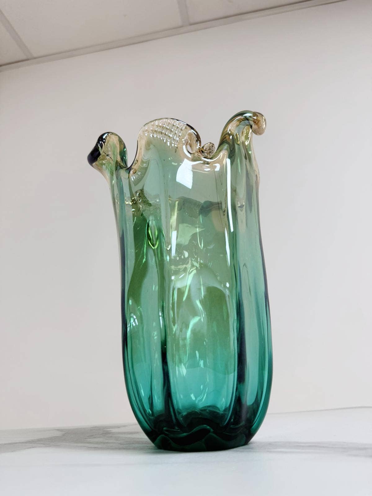Eine mittelgroße, grüne Murano-Glasvase „Venezia“ von Murano Dream (H 25 cm, Ø 14 cm) mit gewelltem Rand und dezenten goldenen Akzenten steht auf weißem Untergrund vor einem schlichten Hintergrund.