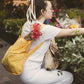 Eine Frau in Weiß mit einer Notabag | „Original Golden“ – einer Kombination aus Tasche und Rucksack – kniet auf einem Blumenmarkt im Freien, hält verpackte Blumen in den Händen und greift nach einem Strauß inmitten üppig grüner Pflanzen.