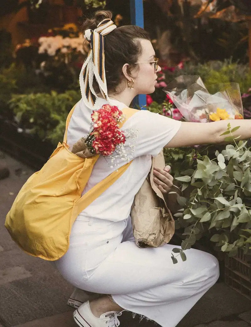 Eine Frau in Weiß mit einer Notabag | „Original Golden“ – einer Kombination aus Tasche und Rucksack – kniet auf einem Blumenmarkt im Freien, hält verpackte Blumen in den Händen und greift nach einem Strauß inmitten üppig grüner Pflanzen.