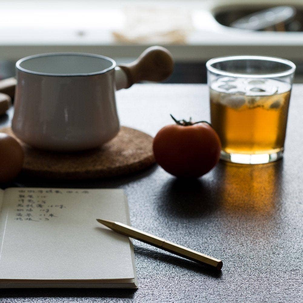Ein ENTREPÔT YSTUDIO Klassischer Revolve Kugelschreiber Schlank liegt auf einem Notizbuch auf einem Tisch neben einer Tomate, Eistee, Eiern und einem weißen Topf mit Holzgriff, der vom Tageslicht sanft beleuchtet wird.