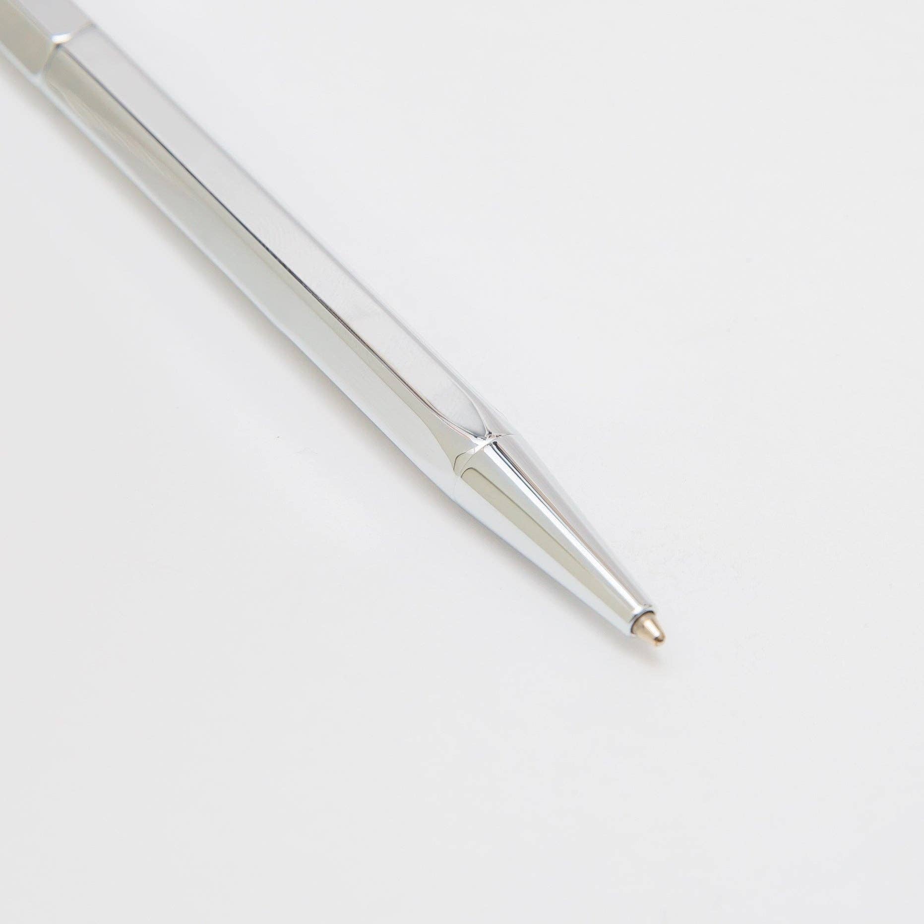Die Nahaufnahme des ENTREPÔT YSTUDIO Klassischer Revolve Kugelschreiber Slim (Glänzendes Silber) zeigt den glatten silbernen Metallkörper und die spitze Spitze, die auf einer weißen Oberfläche ruht.