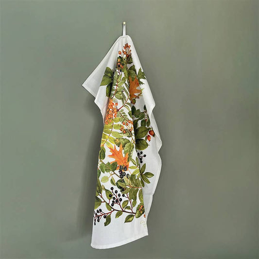 Ein Geschirrtuch von Koustrupco aus 100 % Bio-Baumwolle mit einem herbstlichen Motiv aus bunten Blättern und Beeren hängt an einem Haken vor einer schlichten grünen Wand.