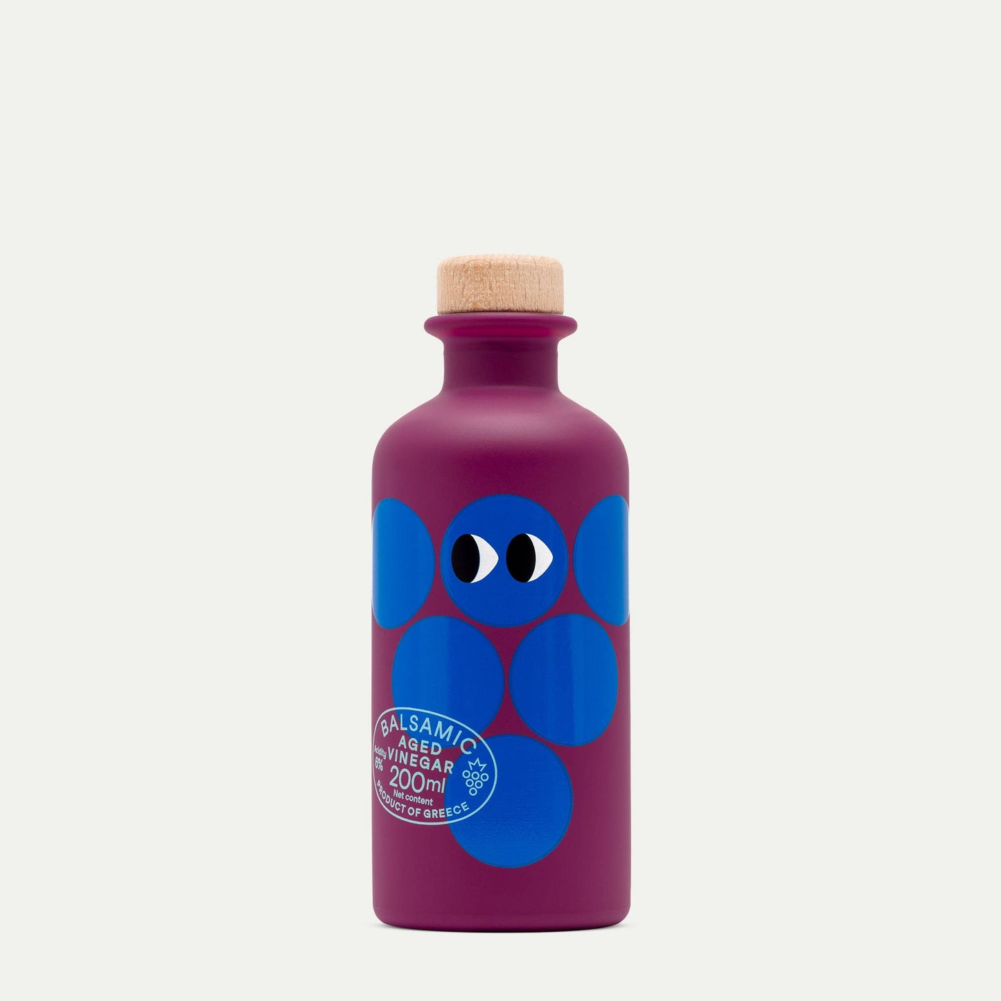 Eine violette Flasche mit einem Holzkorken, blauen Kreisen, zwei Cartoon-Augen und dem Etikett "Yiayia and friends | Gereifter Balsamico-Essig 200 ml," zehn Jahre gereift, Product of Greece by yiayia and friends.