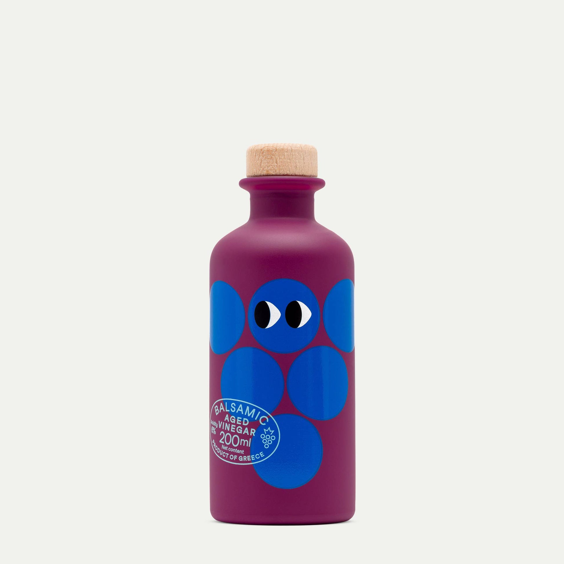 Eine violette Flasche mit einem Holzkorken, blauen Kreisen, zwei Cartoon-Augen und dem Etikett "Yiayia and friends | Gereifter Balsamico-Essig 200 ml," zehn Jahre gereift, Product of Greece by yiayia and friends.