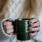 Eine Person mit langen, hellbraunen Haaren in einem weißen Strickpullover hält beide Teile des dunkelgrünen, gerippten Sagaform | Kaffeebecher Grün, 2-tlg von Sagaform GmbH. Ihre Fingernägel sind passend zur Farbe der Becher lackiert.