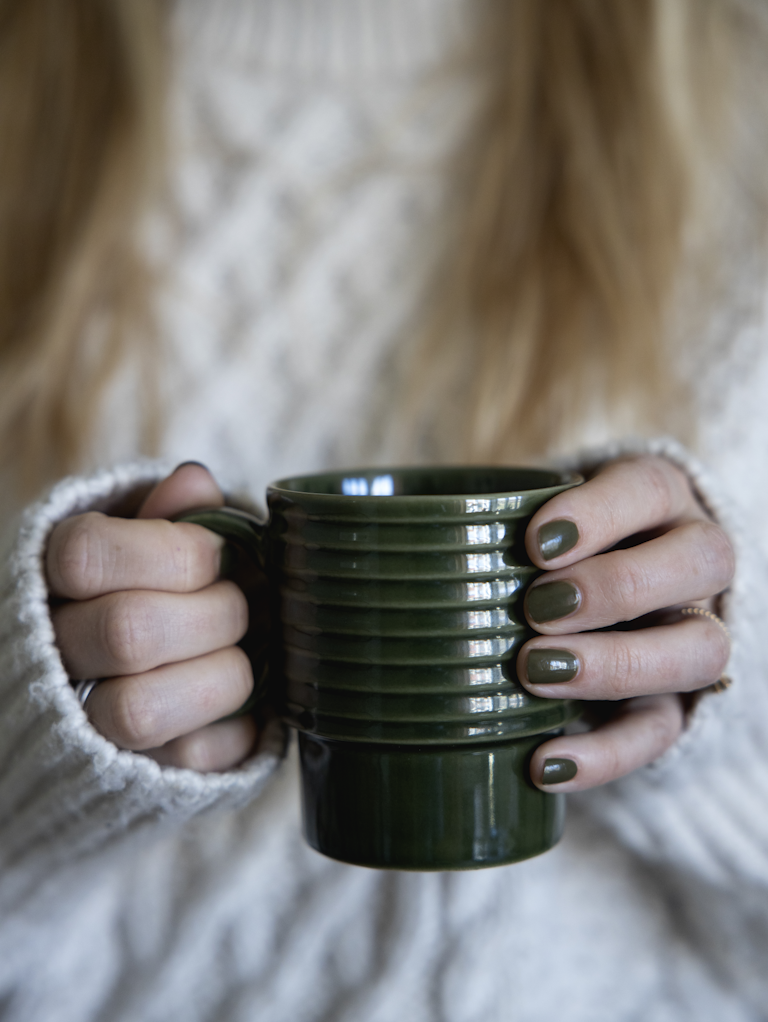 Eine Person mit langen, hellbraunen Haaren in einem weißen Strickpullover hält beide Teile des dunkelgrünen, gerippten Sagaform | Kaffeebecher Grün, 2-tlg von Sagaform GmbH. Ihre Fingernägel sind passend zur Farbe der Becher lackiert.