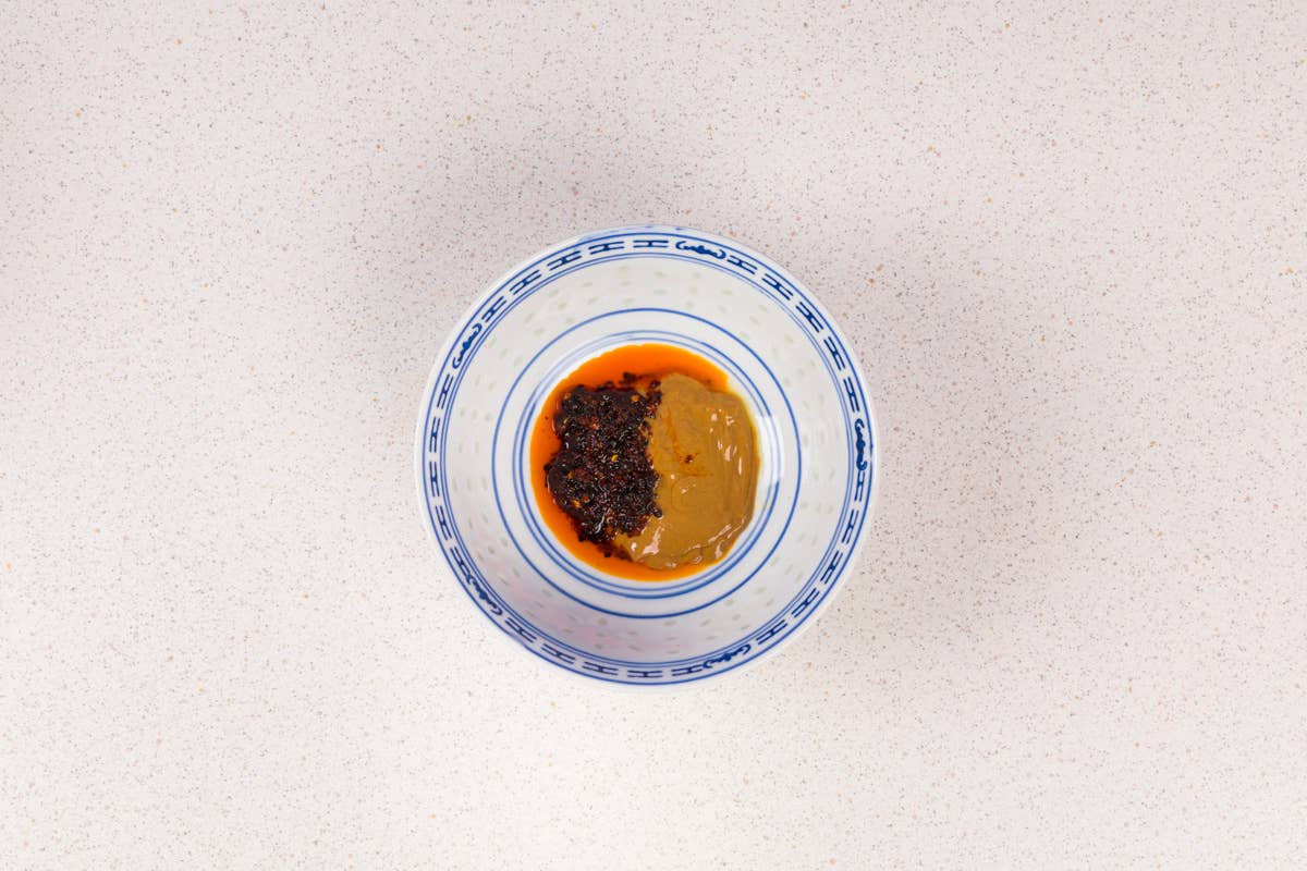 In einer blau-weißen Schüssel befinden sich ein Klecks „White Mausu Ltd White Mausu | Black Bean Rayu Sauce“ – eine braune Paste aus fermentierten Bohnen – und ein Löffel voll scharfer Soße mit Chiliflocken; die Schüssel steht auf einer hell gesprenkelten Arbeitsplatte.