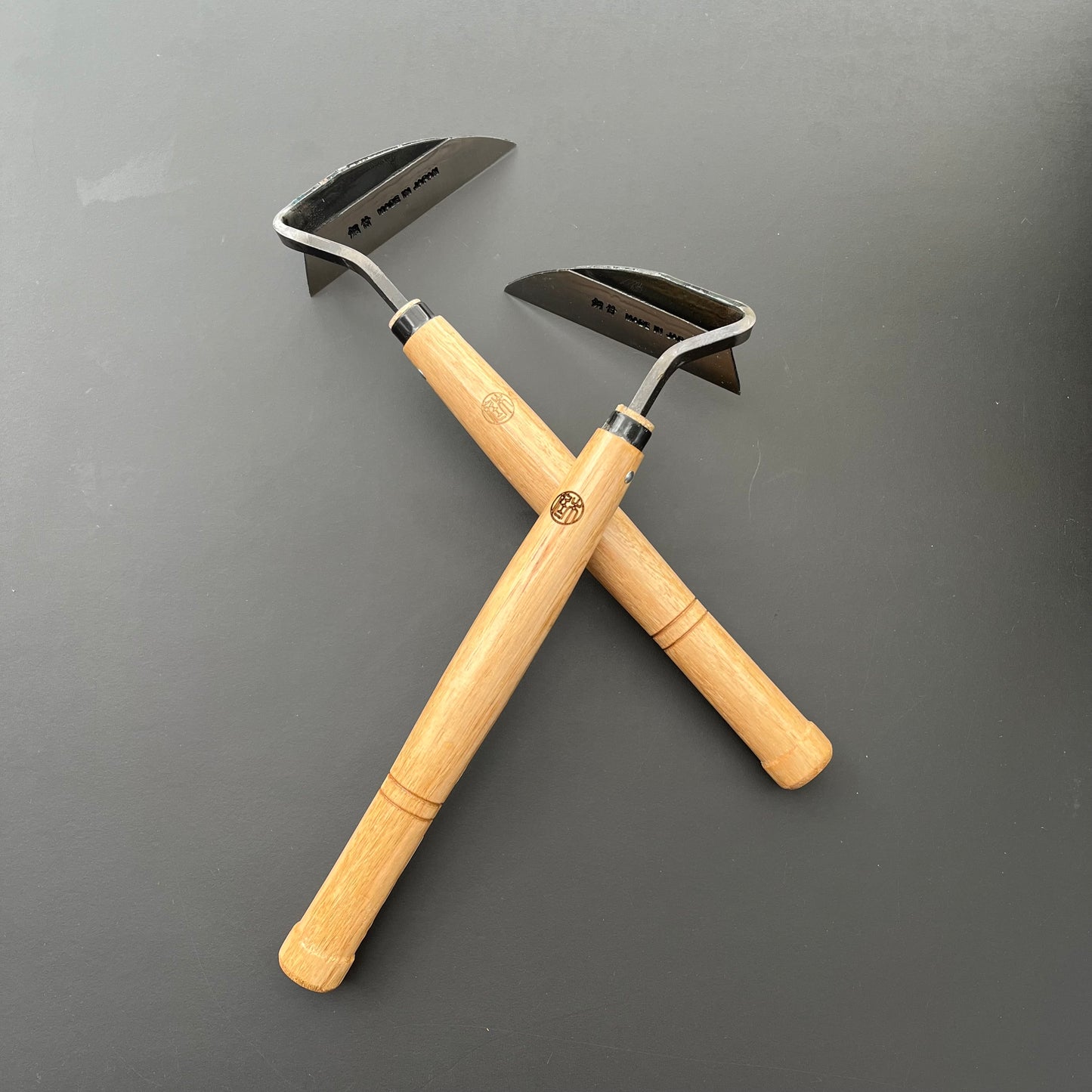 Zwei Niwaki 'Weeding Hoe' Jäthacken für Rechts- und Linkshänder, jeweils mit Holzgriff und abgewinkelter Metallklinge, sind auf einer flachen, dunklen Oberfläche gekreuzt - perfekt zum Entfernen von Unkraut.