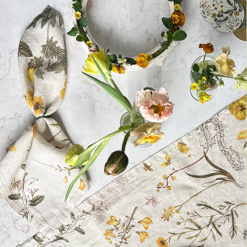 Flat-Lay-Aufnahme eines Tisches, geschmückt mit der „Koustrupco Leinenserviette mit gelben Blütenmotiv“ (45 x 45 cm), gelben und rosa Blumen in kleinen Vasen, einem Blumenkranz und einem Teller – eine bezaubernde botanische Tischdekoration auf hellem Marmor.