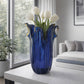 Die „Murano Dream Große Giusy“-Vase aus blauem Original-Murano-Glas (H 32, Ø 20 cm) mit gewellten Rändern, gefüllt mit weißen Tulpen, steht auf einem weißen Tisch in einem modernen Wohnzimmer mit großen Fenstern und einem grauen Sofa.