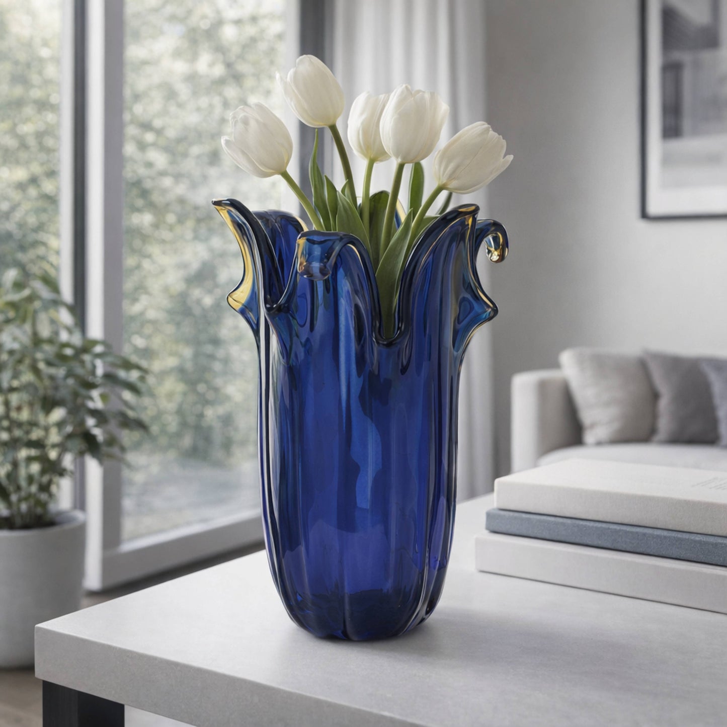 Die „Murano Dream Große Giusy“-Vase aus blauem Original-Murano-Glas (H 32, Ø 20 cm) mit gewellten Rändern, gefüllt mit weißen Tulpen, steht auf einem weißen Tisch in einem modernen Wohnzimmer mit großen Fenstern und einem grauen Sofa.