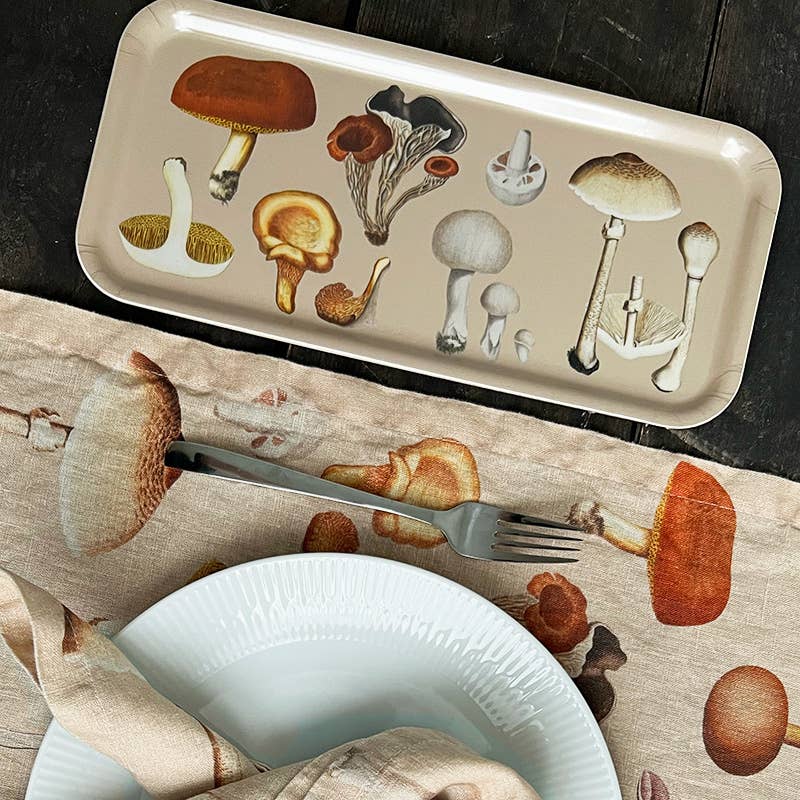 Ein rechteckiges Koustrupco-Serviertablett (32 x 15 cm) mit dem Namen „Pilz“, verziert mit Vintage-Pilzillustrationen, steht auf einem dunklen Tisch neben einem Teller, einer Gabel und einer Tischdecke mit ähnlichen Pilzmotiven und versprüht den zeitlosen Charme eines klassischen „Flora Danica“-Tabletts.