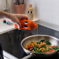 Eine Hand gießt Yiayia and friends | Natives Olivenöl extra mit Chili aus einer orangefarbenen Flasche in eine Pfanne mit Kichererbsen, Spinat und rotem Paprika und verleiht der Küchenszene einen mediterranen Touch.