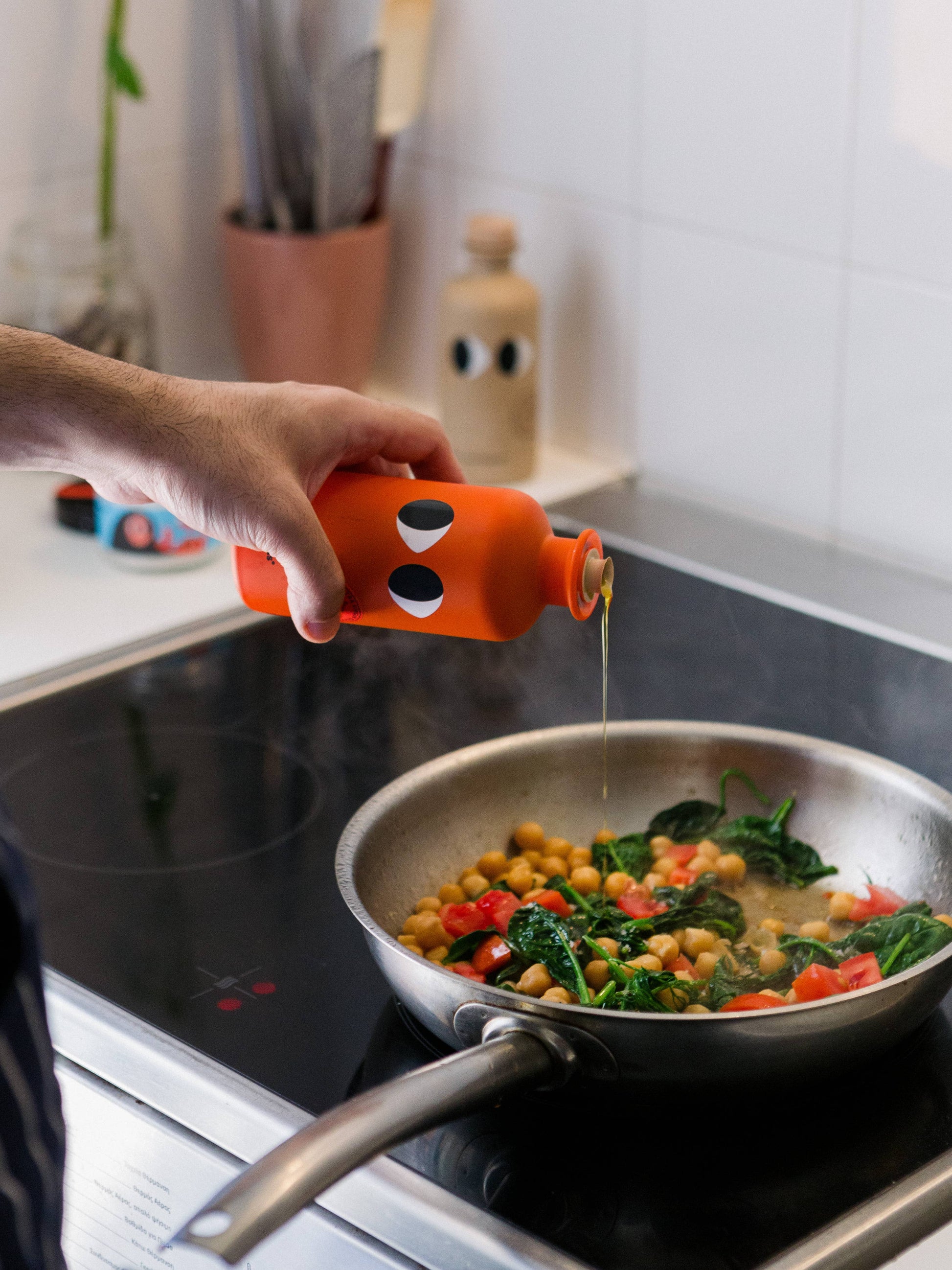 Eine Hand gießt Yiayia and friends | Natives Olivenöl extra mit Chili aus einer orangefarbenen Flasche in eine Pfanne mit Kichererbsen, Spinat und rotem Paprika und verleiht der Küchenszene einen mediterranen Touch.