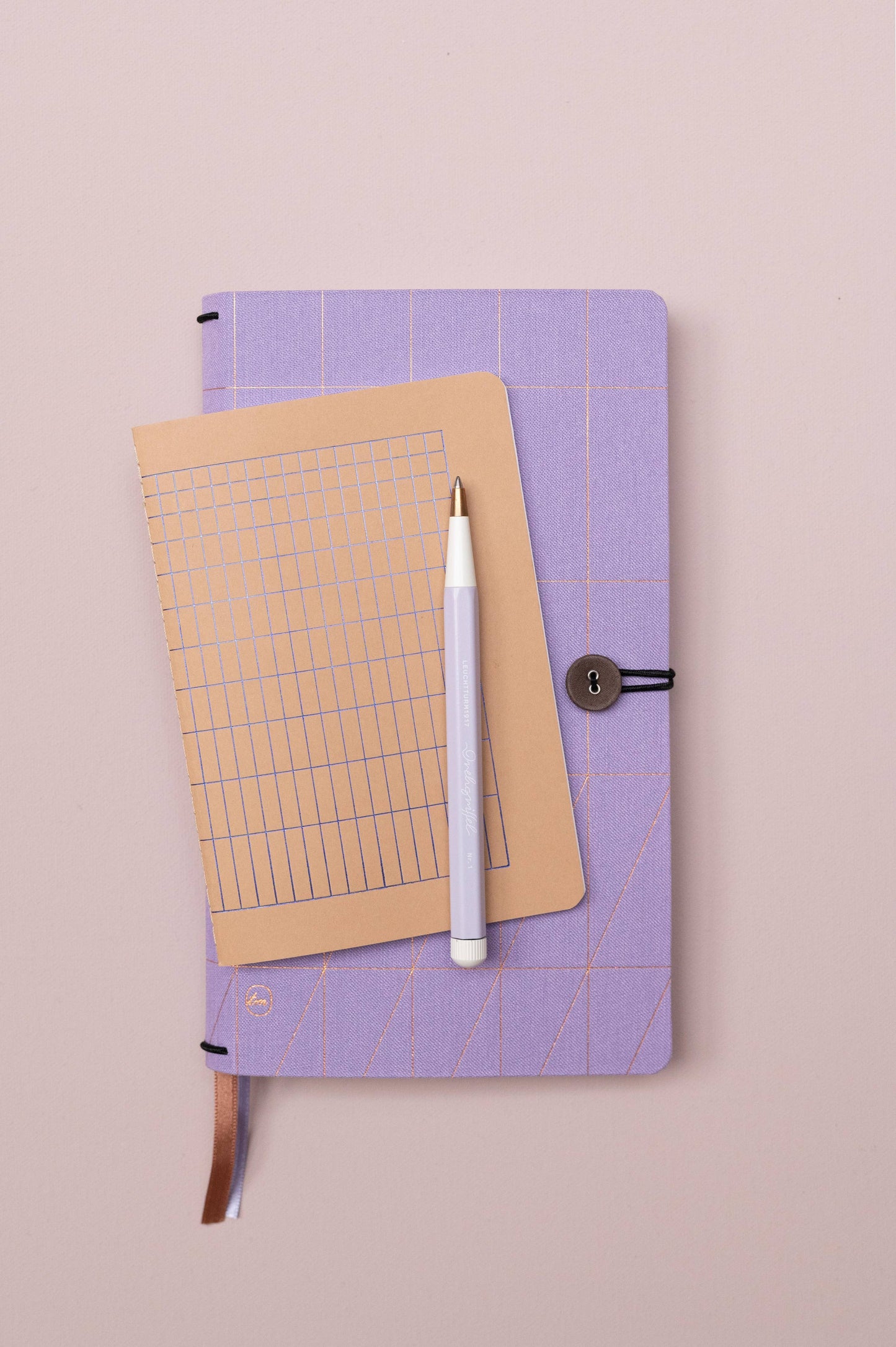 Das Tinne Mia Notizbuch A6 'Nougat Grid' mit FSC-Papier und beige gepunktetem Raster liegt samt weißem Stift auf einem lavendelfarbenen Notizbuch mit Knopfverschluss auf zartrosafarbenem Untergrund.