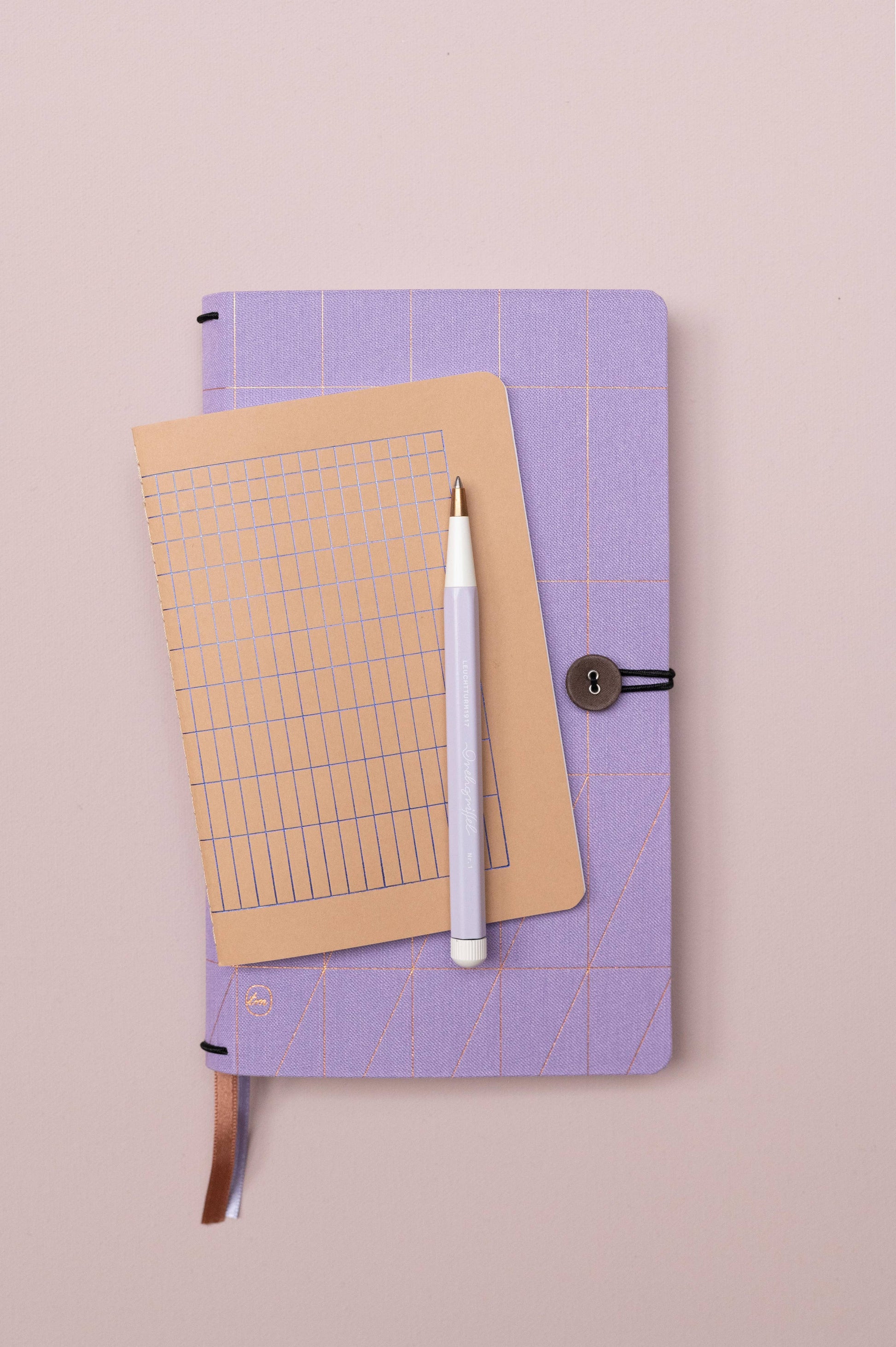 Das Tinne Mia Notizbuch A6 'Nougat Grid' mit FSC-Papier und beige gepunktetem Raster liegt samt weißem Stift auf einem lavendelfarbenen Notizbuch mit Knopfverschluss auf zartrosafarbenem Untergrund.