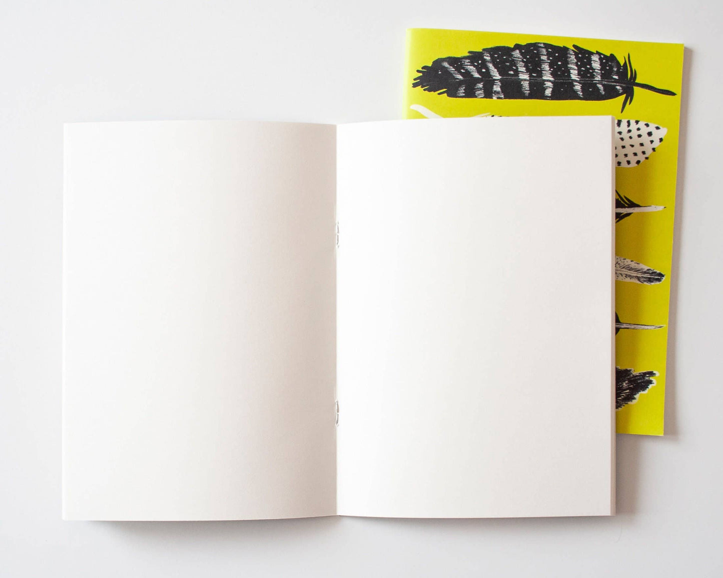 Ein offenes Blanko-Notizheft DIN A5 liegt auf dem geschlossenen Papaya Paper Products | Schönes Notizheft A5 mit Federn in gelb, beide aus FSC-zertifiziertem Munken Print Cream Papier, angeordnet auf einer weißen Fläche.