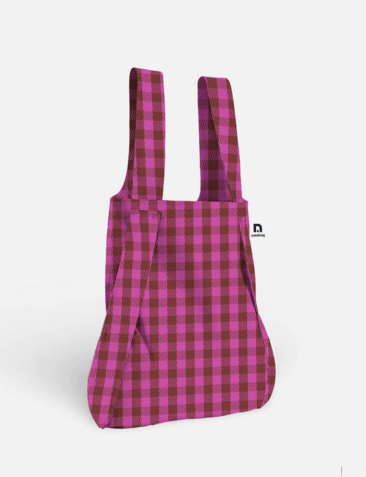Die Notabag | Wandelbare Tasche und Rucksack „Kirsche“ von Notabag steht aufrecht vor einem schlichten weißen Hintergrund und präsentiert ihr rosa-burgunderrotes Karomuster. Diese innovative Tasche lässt sich mühelos von einer Tragetasche in einen Rucksack verwandeln – eine wahrhaft wandelbare Tasche für den Alltag.