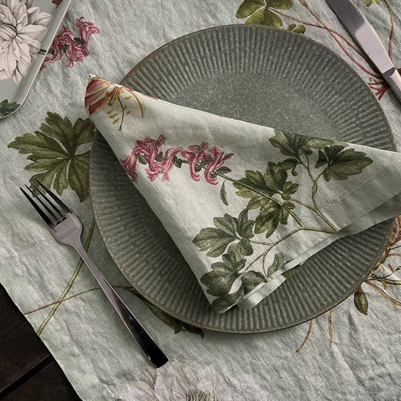 Ein strukturierter grüner Teller, auf dem die Leinserviette „Flora Danica Atlas“ von Koustrupco (45 × 45 cm) gefaltet liegt, auf einer passenden Tischdecke mit Blumenmuster. Zu beiden Seiten des Tellers liegen eine Gabel und ein Messer.
