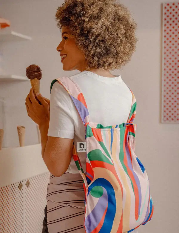 Eine lächelnde Person mit lockigem Haar hält eine Schokoladeneiswaffel und trägt die Notabag 'Flow' Kombination aus Tasche & Rucksack von Notabag über der Schulter, während sie in einem Innenraum neben leeren Eistüten auf einer Theke steht.