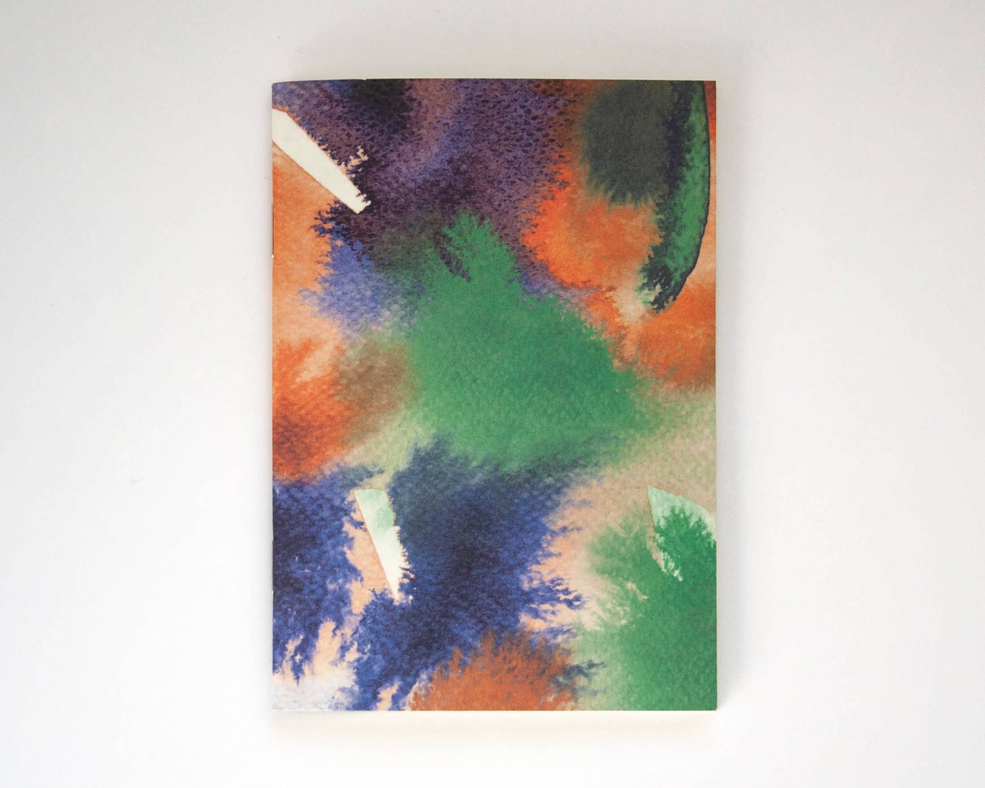 Das Papaya Paper Products | Notizheft A6 mit Aquarell/Watercolor-Motiv, FSC zertifiziert und mit blanko Seiten, zeigt bunte grüne, orange, lila und blaue Flecken auf dem Cover und liegt geschlossen auf weißem Hintergrund.