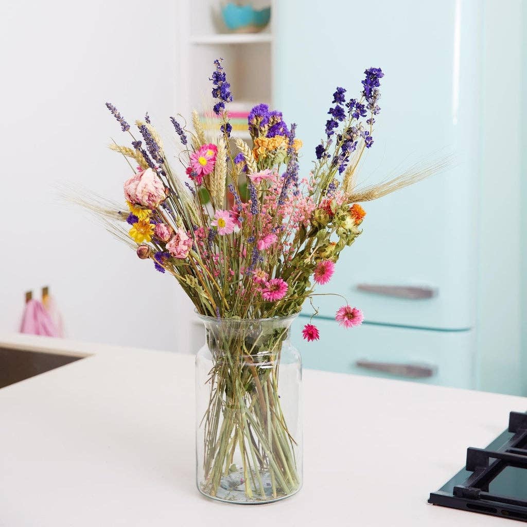 In einer Vase aus klarem Glas sind die farbenfrohen Trockenblumen „Feldblumenstrauß“ S – M von Wildflowers by Floriette zu sehen, die zusammen mit bunten getrockneten Wildblumen auf einer weißen Küchenarbeitsplatte stehen, im Hintergrund ist ein hellblauer Kühlschrank zu erkennen.