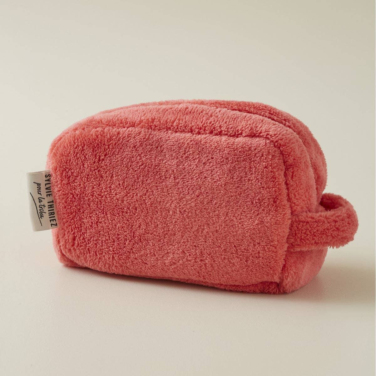 Eine weiche, rechteckige rosa Sylvie Thiriez Kosmetiktasche Flamingofarben aus Plüsch mit kleinem Henkel und einem "SMILE THERE!"-Etikett ruht auf einer schlichten, hellen Fläche.